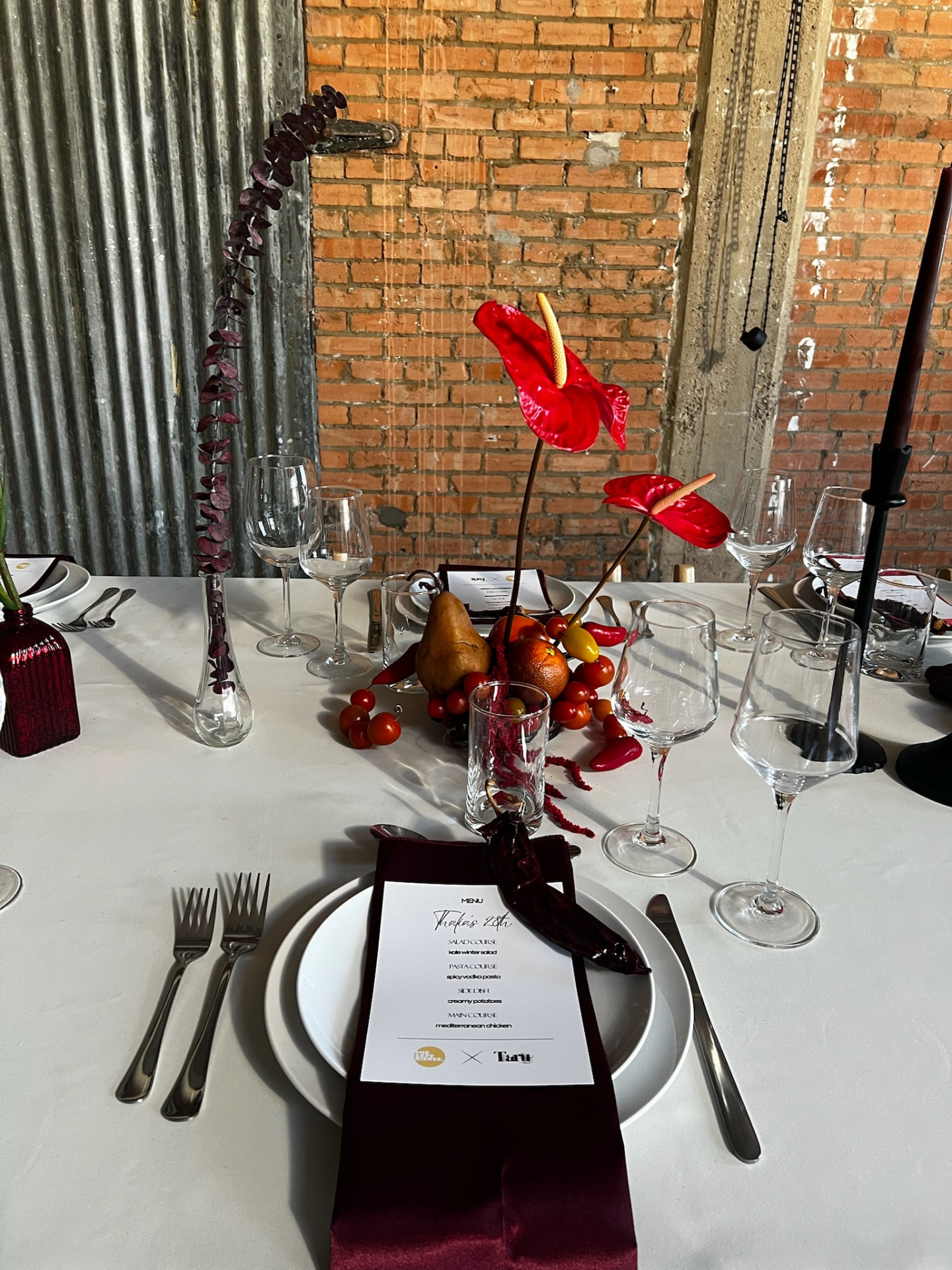 Close-up place setting beside red anthurium and a cluster of fruit on a set dinner table