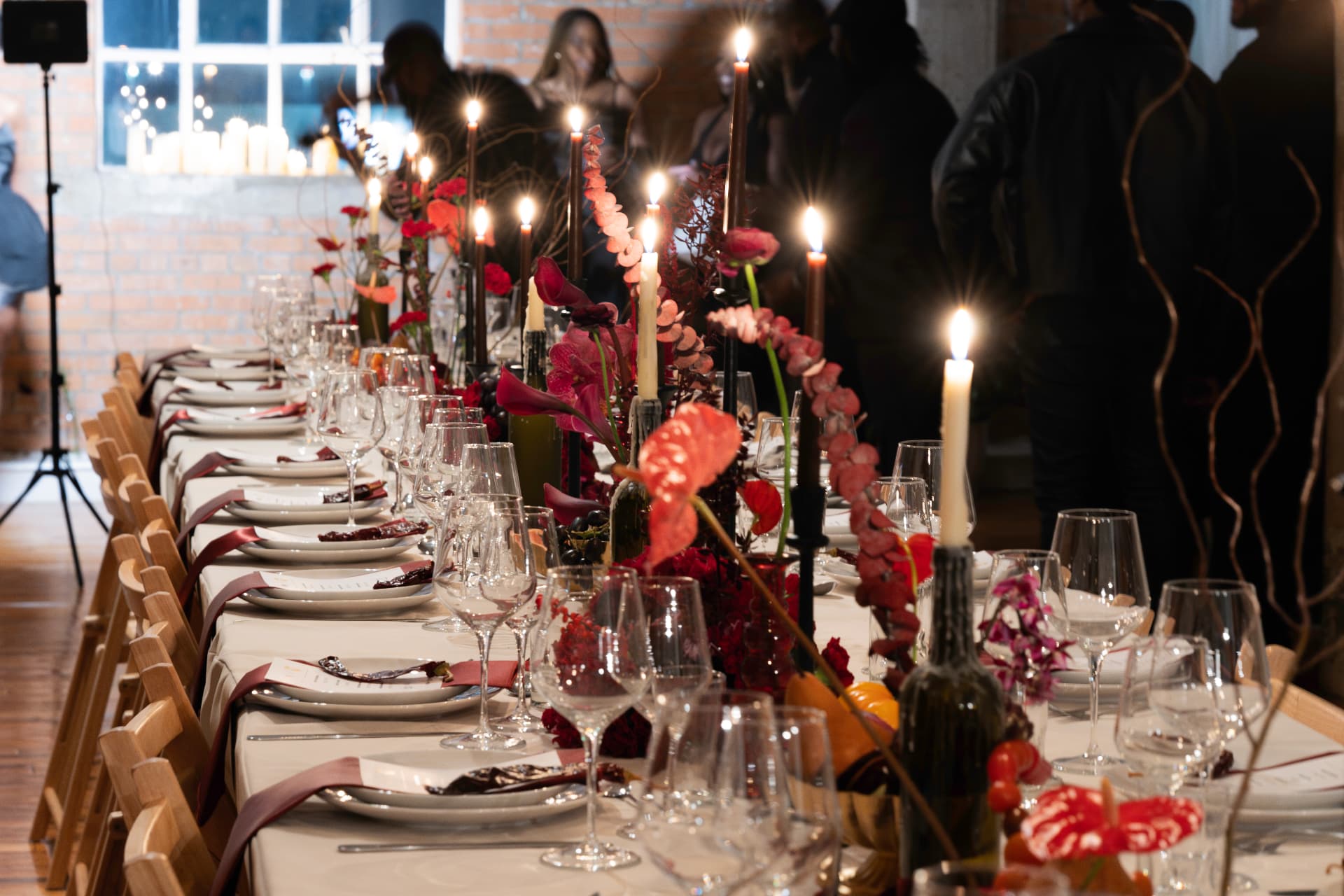 Long candlelit dinner table with red florals and tapers in a brick-walled room