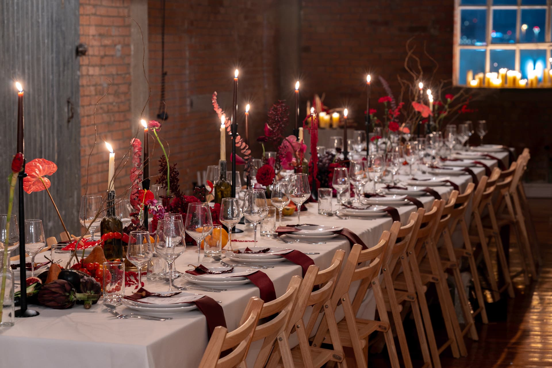 Full view of a long banquet table lined with chairs, red centrepieces, and candlelight against a brick wall